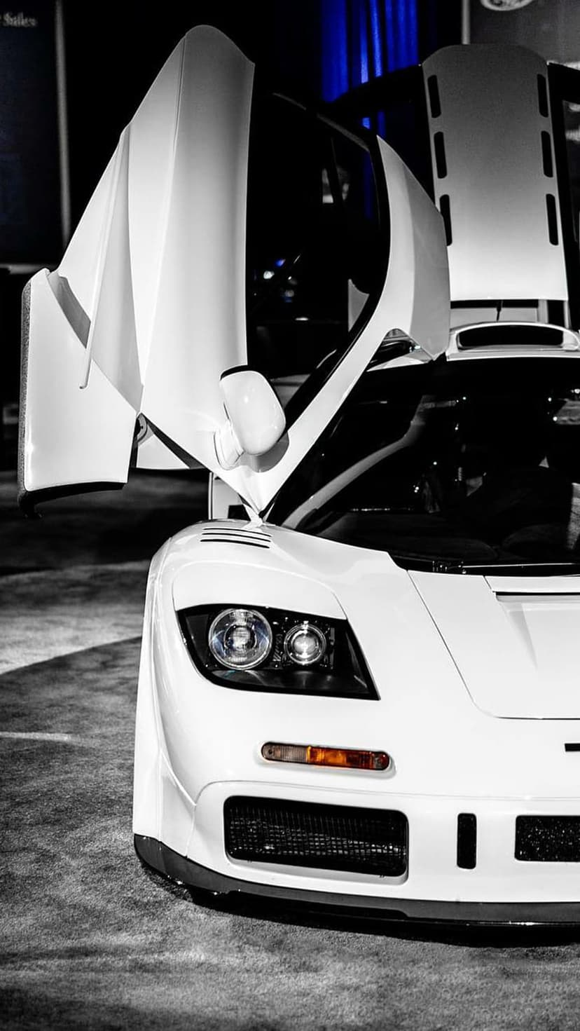 White McLaren F1 supercar with open butterfly doors and round headlights shown in a black and white low-angle view.