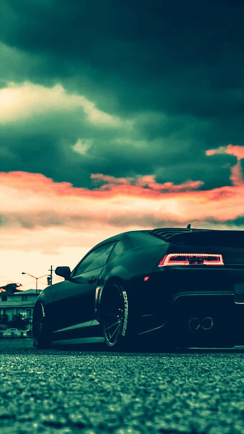 Black Camaro rear view on asphalt under a dramatic orange sunset sky with dark moody clouds and glowing exhaust.