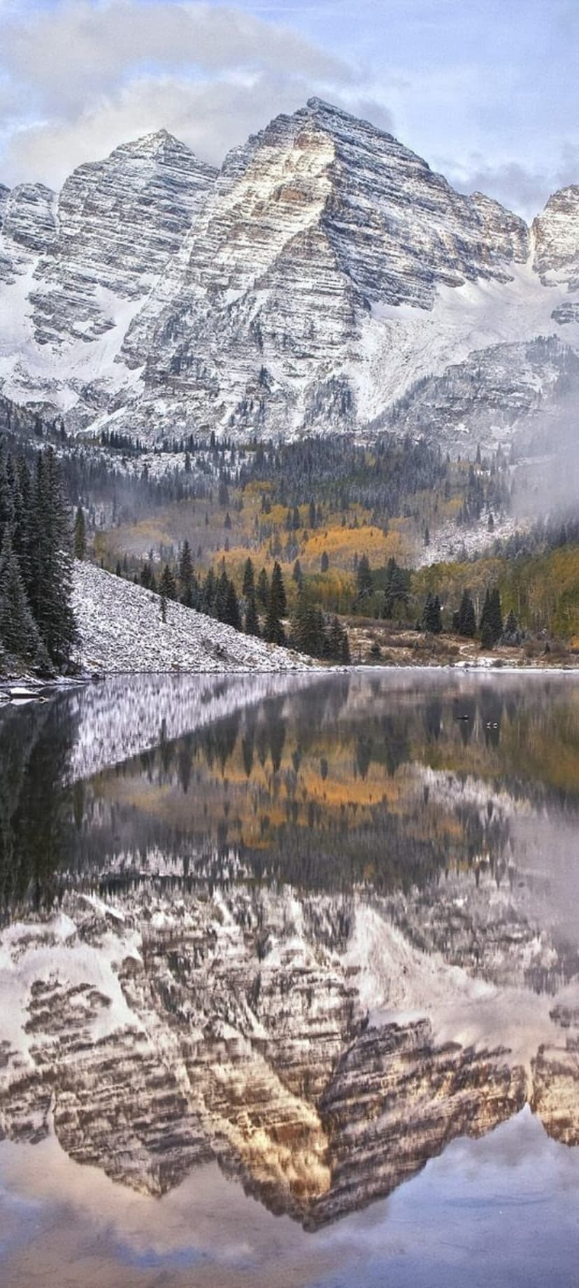 Snow-covered Maroon Bells peaks reflecting in a calm lake surrounded by yellow autumn aspens and evergreens.