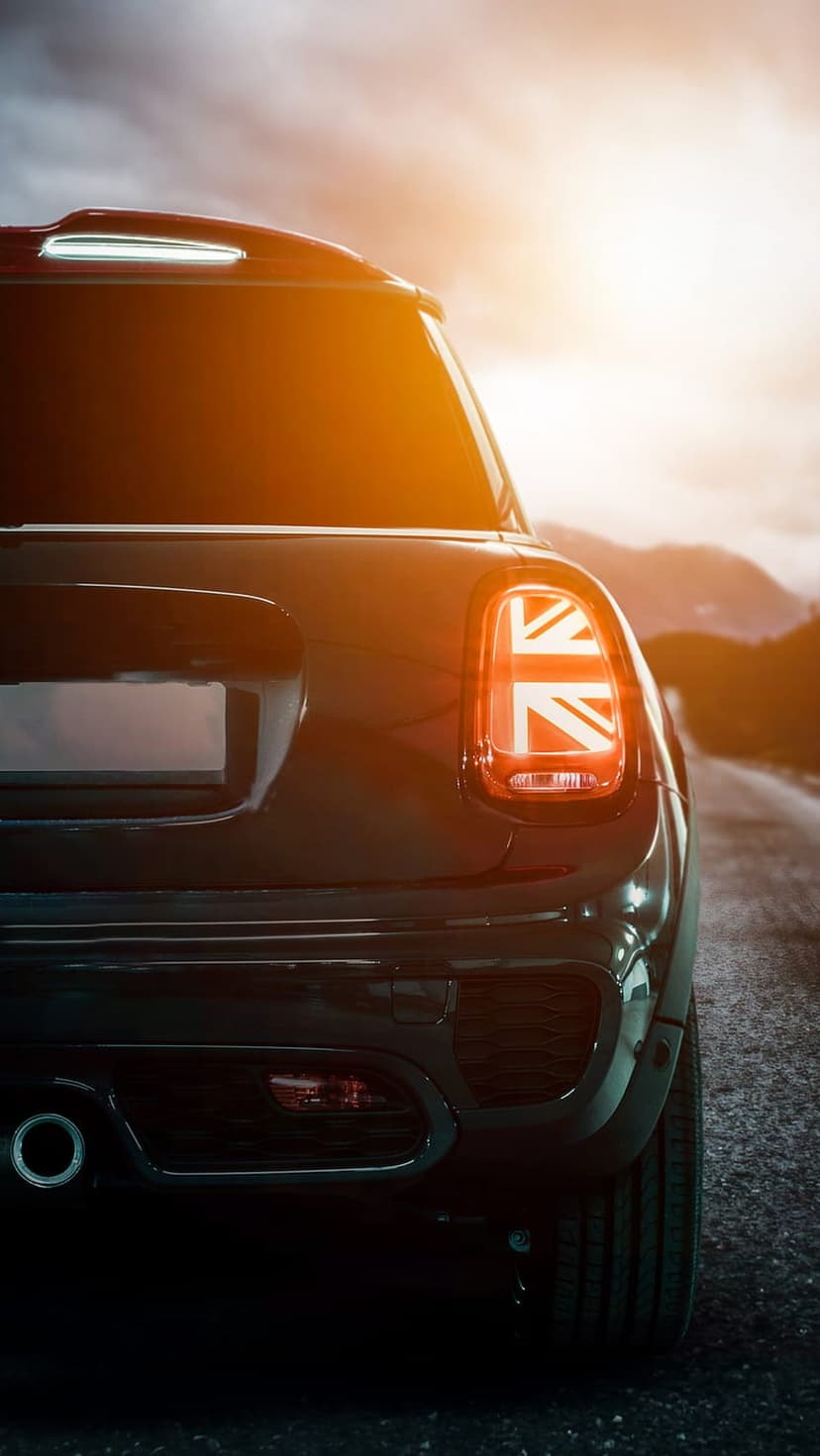 Mini Cooper taillight with a Union Jack design glows red against a sunset mountain silhouette and golden sky.