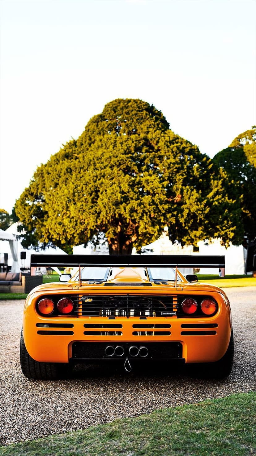 Orange McLaren F1 GTR race car with a large rear wing and quad exhausts parked on a gravel path near a tree.