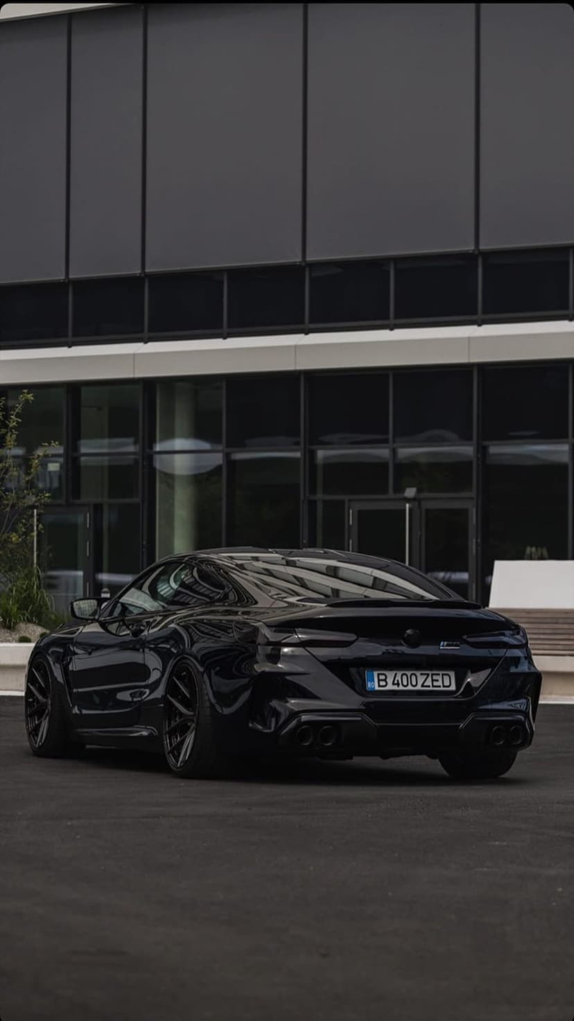 Black sports car with widebody fenders parked on asphalt in front of a modern glass building at night.