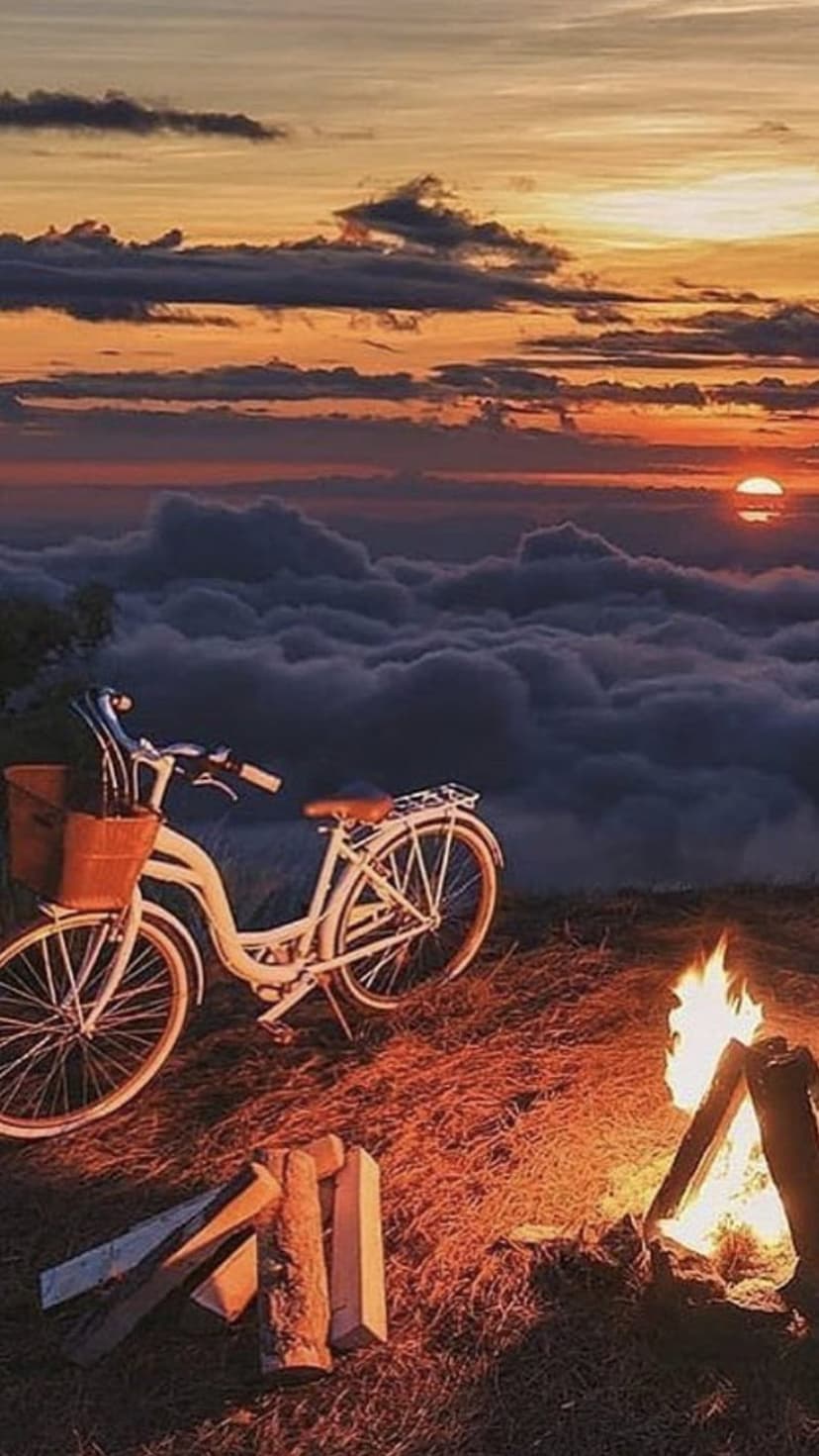 White bicycle with a basket stands near a burning campfire on a grassy hill overlooking a thick layer of clouds.