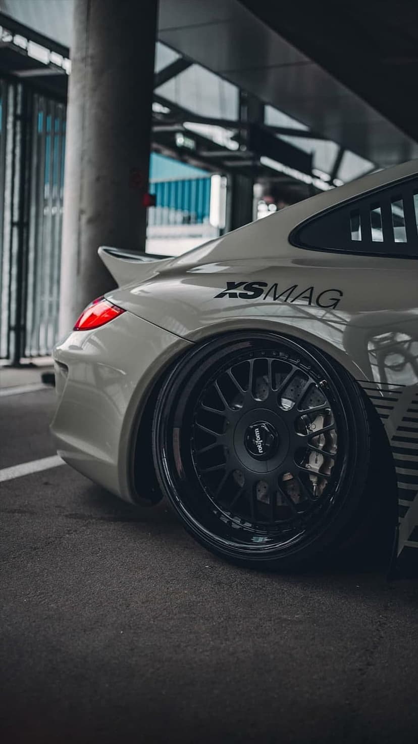 White Porsche rear quarter panel showing custom black multi-spoke wheels, a red taillight, and XSMAG decal.