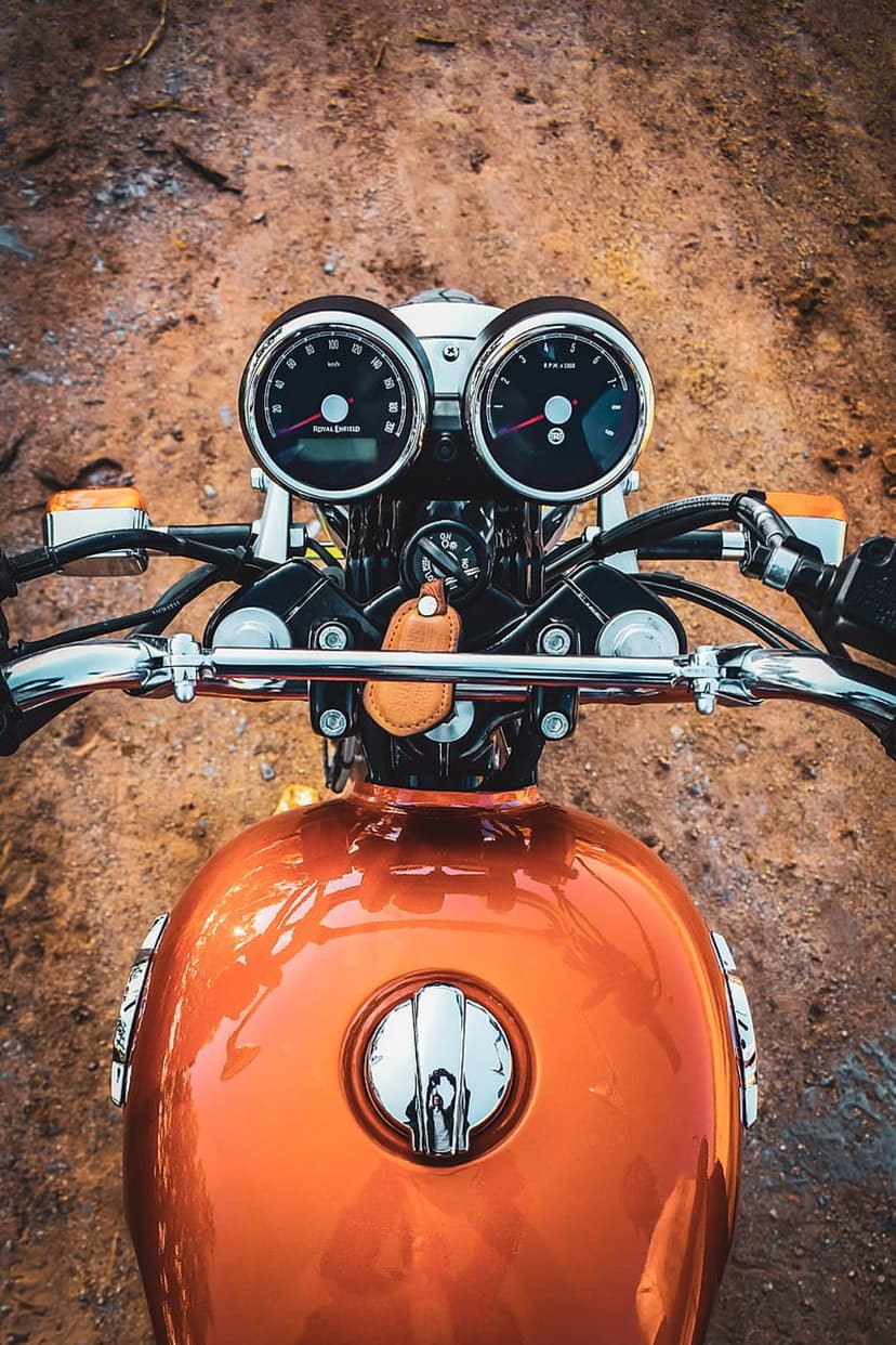 Orange motorcycle fuel tank and dual chrome gauges seen from a top-down perspective against a rustic background.