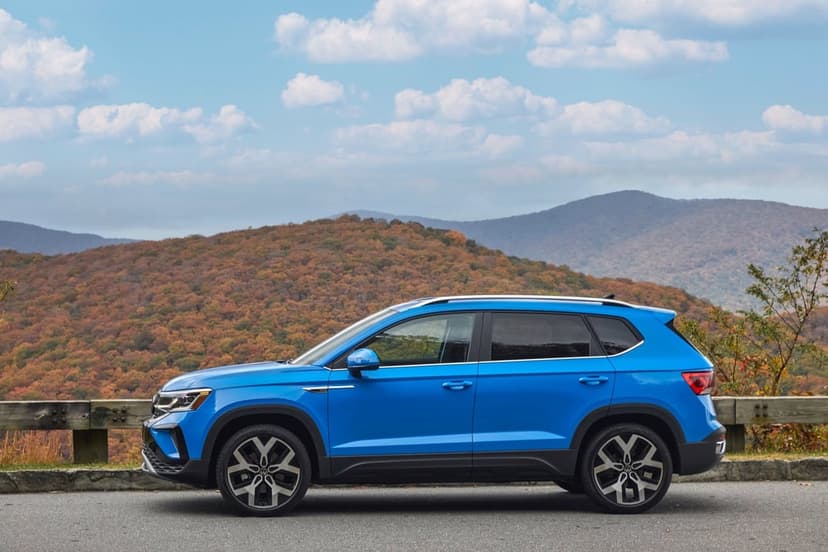 Blue SUV parked on a roadside overlook with rolling autumn-colored mountains and a cloudy blue sky background.