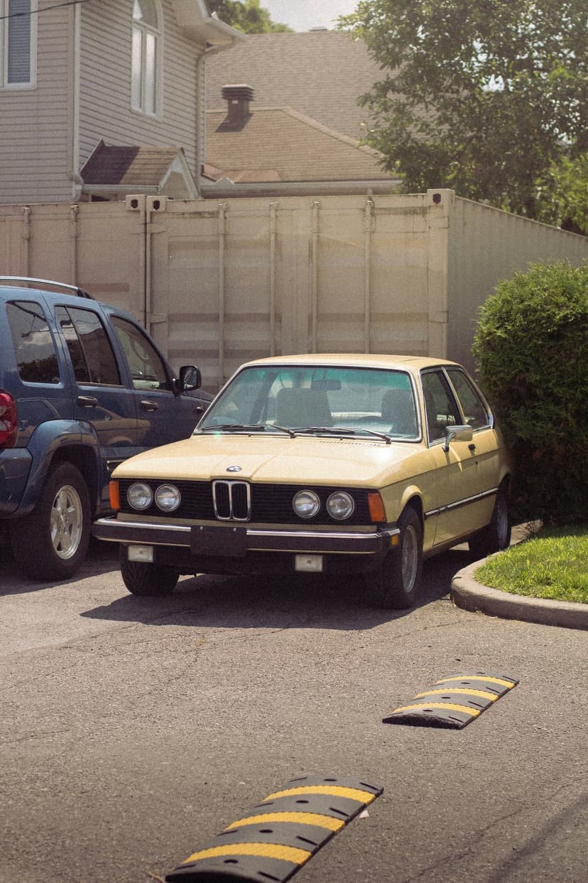 Yellow BMW E30 sedan parked next to a blue SUV in a lot with striped speed bumps on the asphalt ground.
