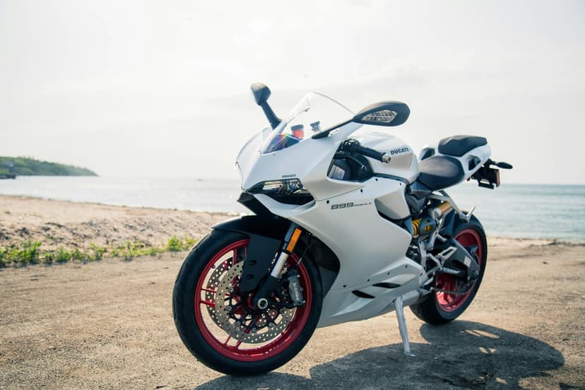 White Ducati motorcycle with red rims parked on a sandy beach next to calm ocean waves under a bright sunny sky.