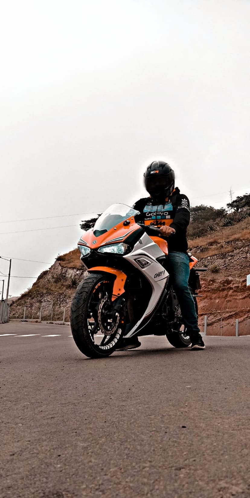 Motorcyclist in a helmet rides an orange and white sportbike on an asphalt road with green hills in the background.