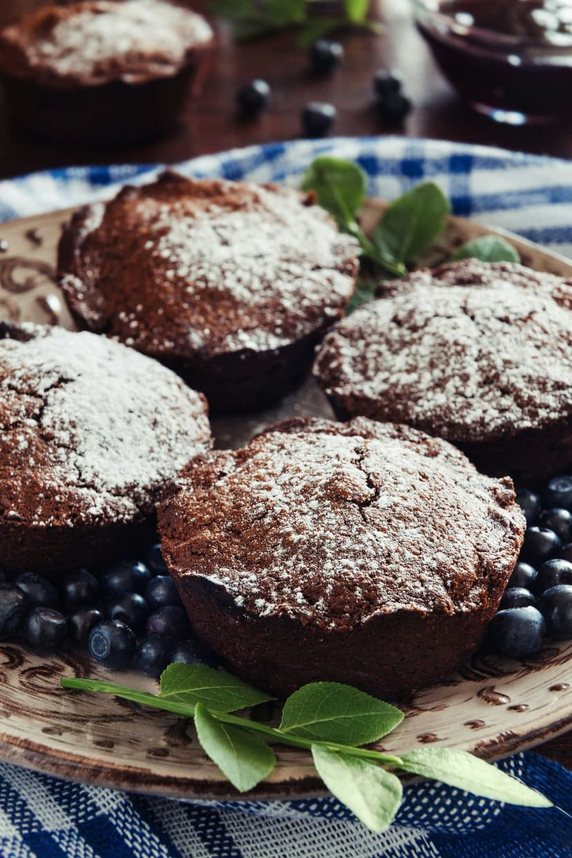 Chocolate muffins dusted with powdered sugar, fresh blueberries, and mint on a rustic plate for a mobile screen.