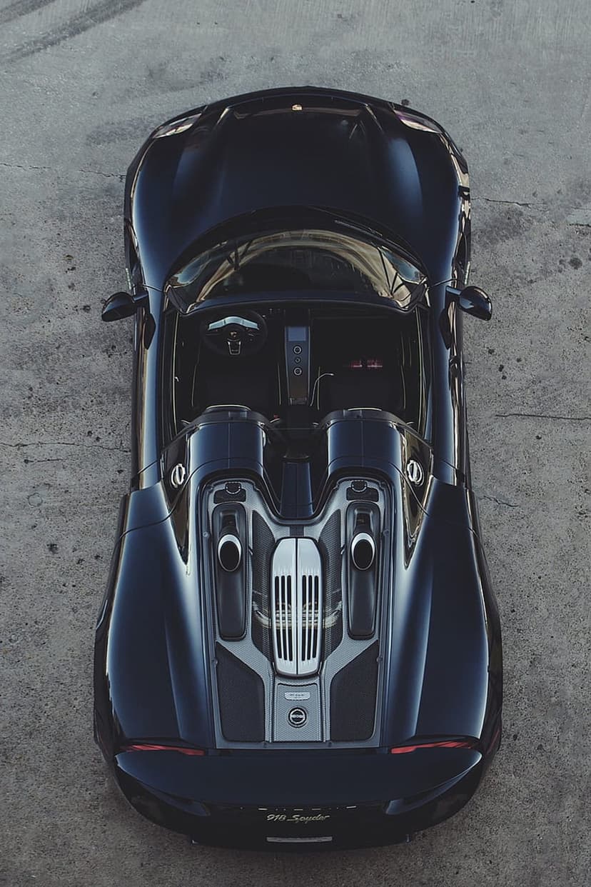 Porsche 918 Spyder in black seen from above on a gray concrete floor with visible dual exhaust pipes.