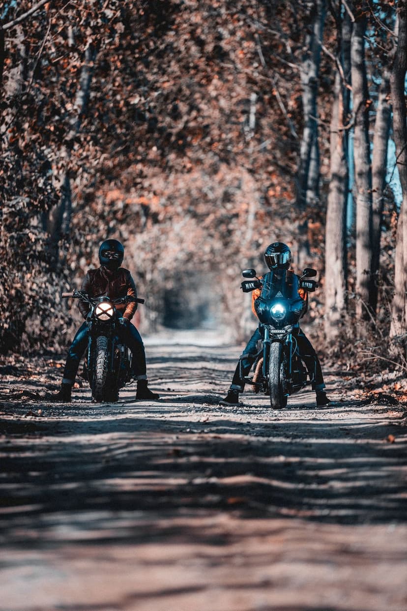 Two motorcyclists in gear stand on a dirt path covered in fallen autumn leaves under a sun-drenched forest canopy.
