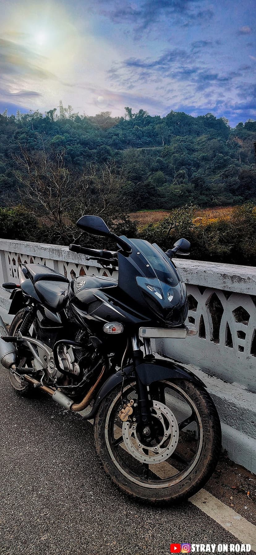 Black motorcycle parked on a bridge overlooking verdant mountain ridges under a heavy grey overcast sky.