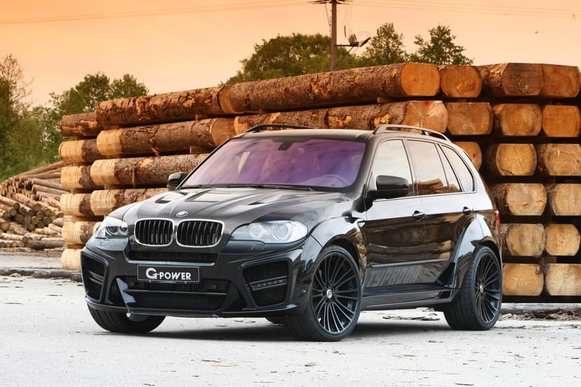 Black BMW X5 M Typhoon SUV with a widebody kit parked in front of large wooden logs during a golden sunset.