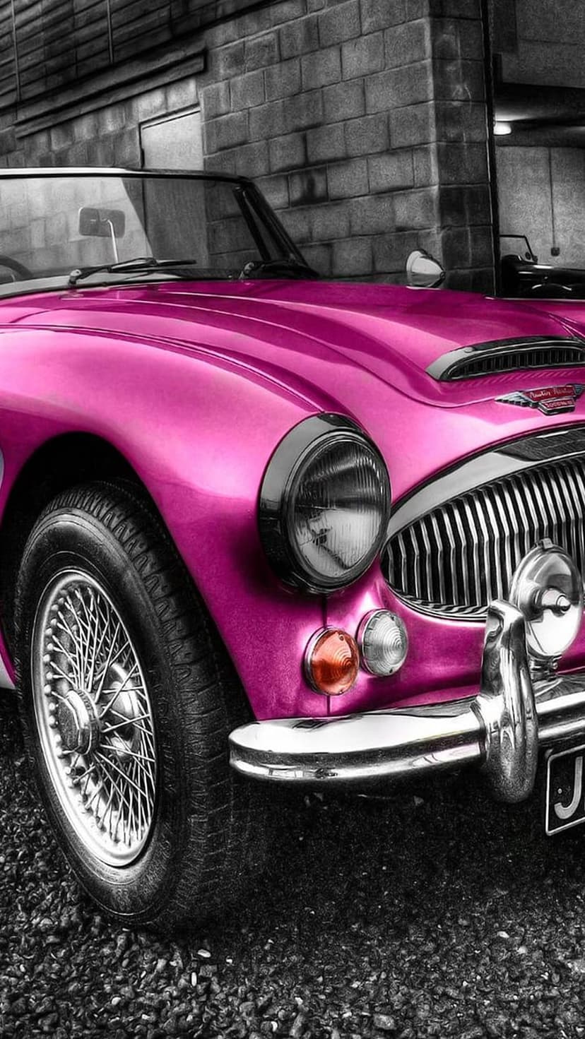 Pink classic car featuring chrome bumpers and wire wheels parked against a gray brick wall on gravel.