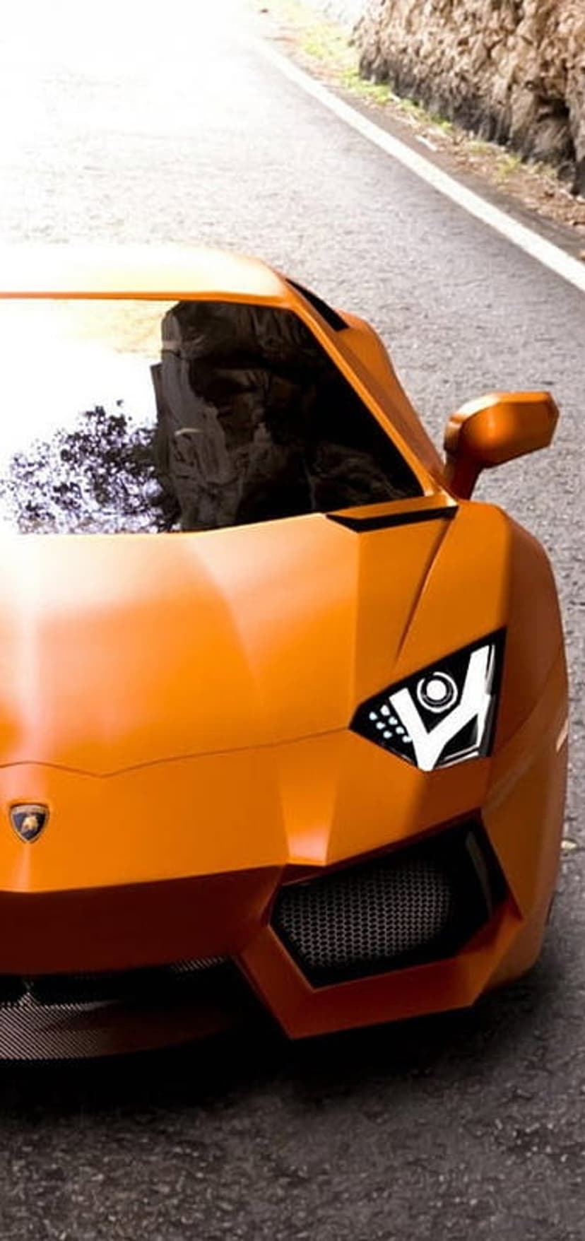 Orange Lamborghini Aventador with glowing LED headlights drives on a paved road during a scenic daytime commute.