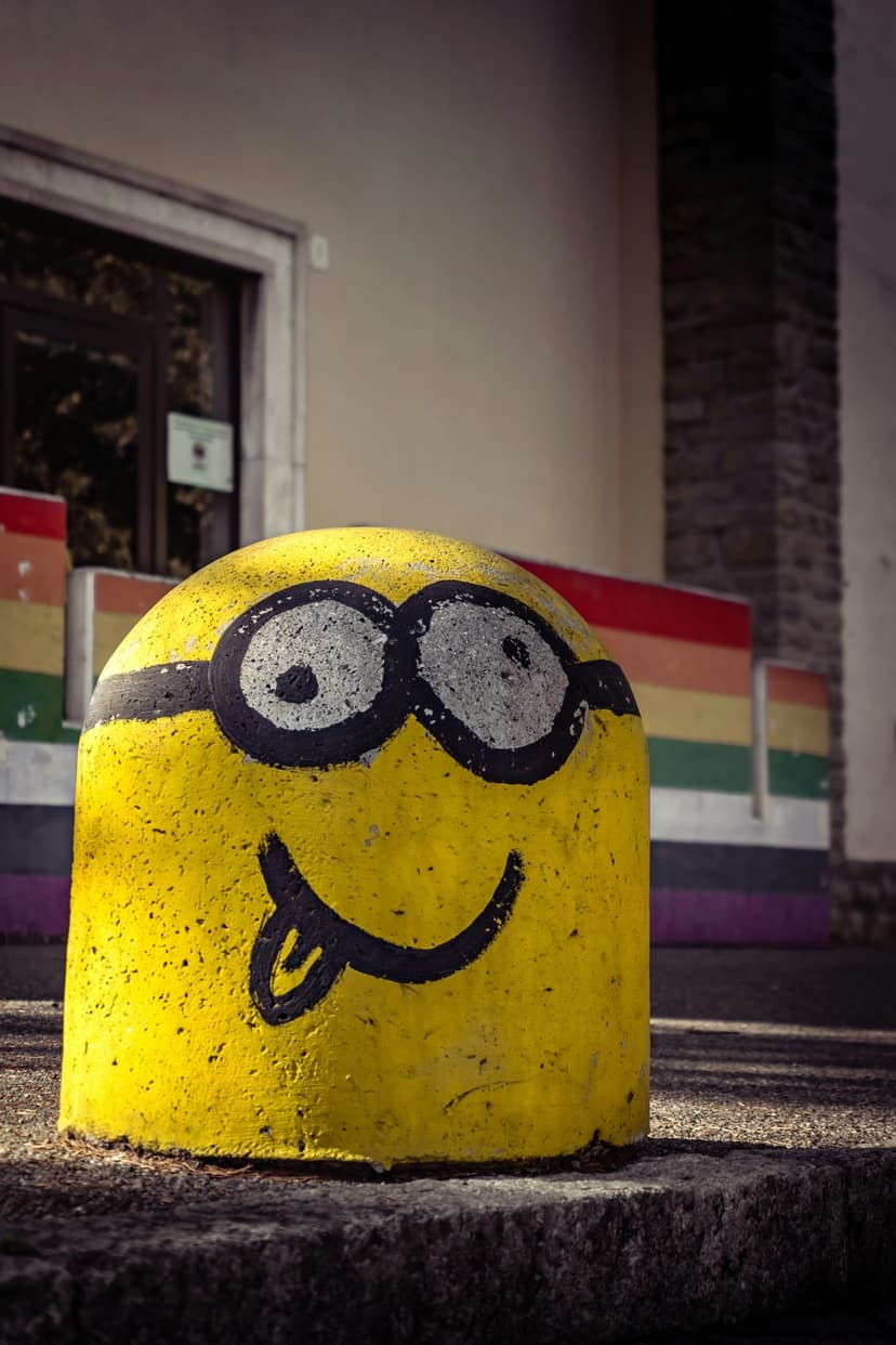 Yellow Minion painted on a concrete bollard with a tongue out and goggles against rainbow panels in sunlight.