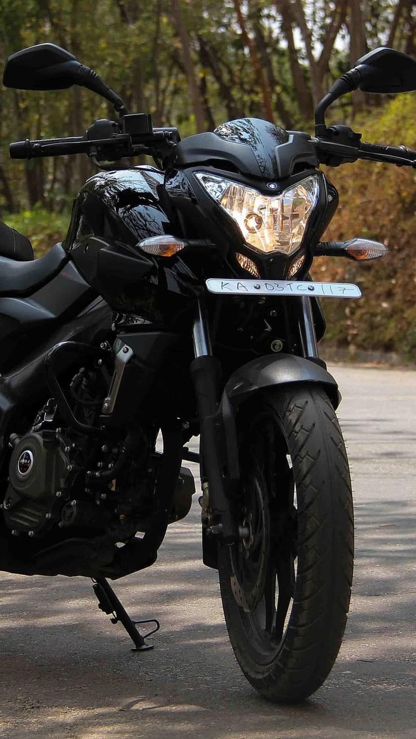 Black sportbike with dual headlights and exposed engine parts parked against a soft-focus forest background.