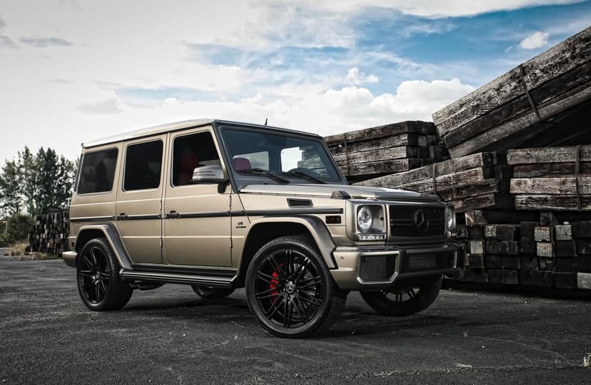 Matte gold Mercedes-Benz G-Wagon parked on asphalt next to stacks of wooden beams under a cloudy sky.
