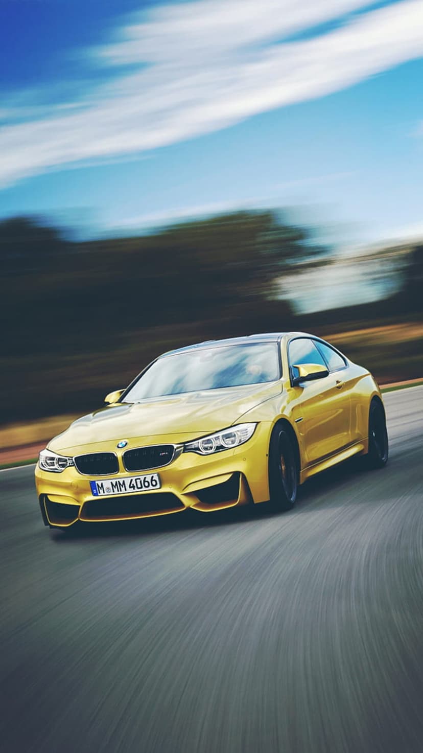 Yellow BMW M4 Coupe speeds on a race track with motion-blurred trees and blue sky in a vertical orientation.