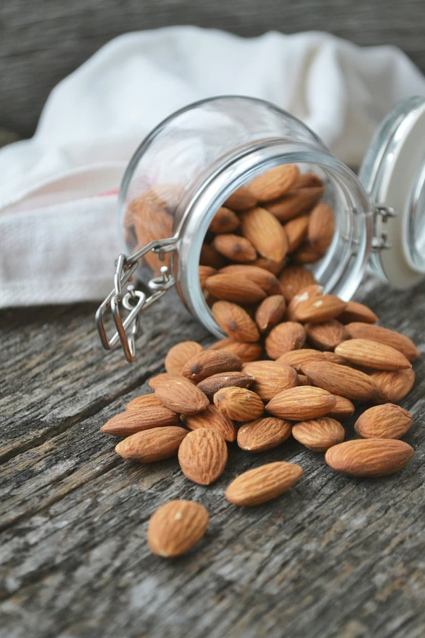 Raw almonds tumble out of a clear glass jar onto a rustic wooden surface with deep grain and warm lighting.