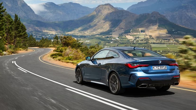Blue BMW 4 Series Coupé driving on a winding mountain road with rugged peaks and valley views.