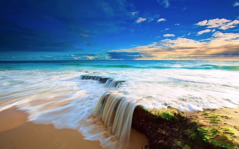 Cascading waterfall spills over emerald mossy rocks on a sandy beach during a vibrant golden hour sunset.