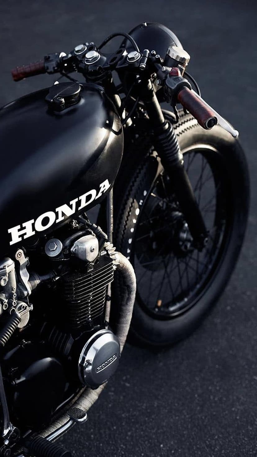 Honda cafe racer fuel tank with white logo, ribbed engine block, and brown leather handlebar grips in dark light.