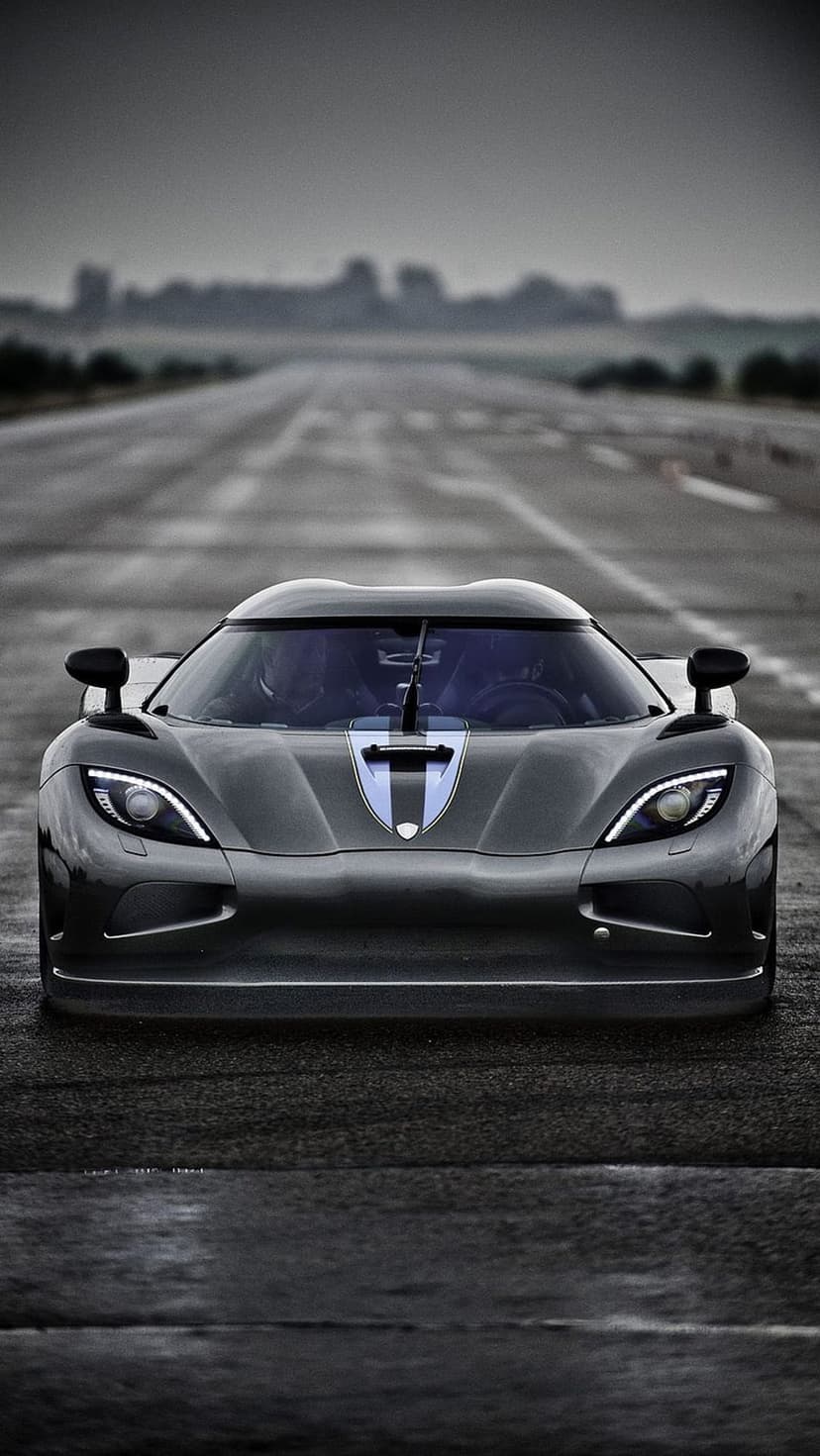 Koenigsegg Agera RS hypercar front view on wet asphalt with glowing headlights and a dark moody sky background.