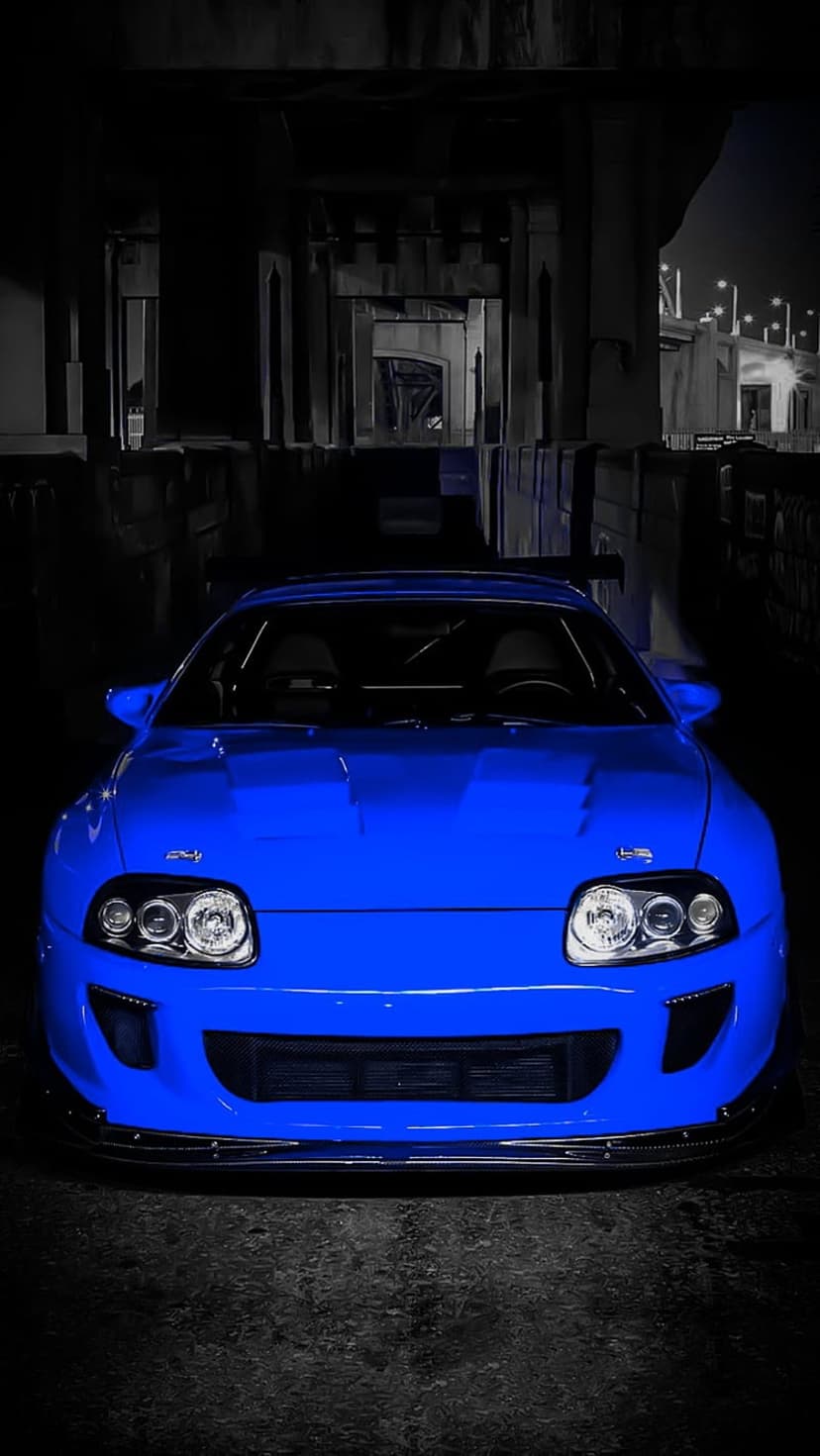 Blue Toyota Supra with glowing headlights parked in a dark industrial underpass from a low-angle perspective.