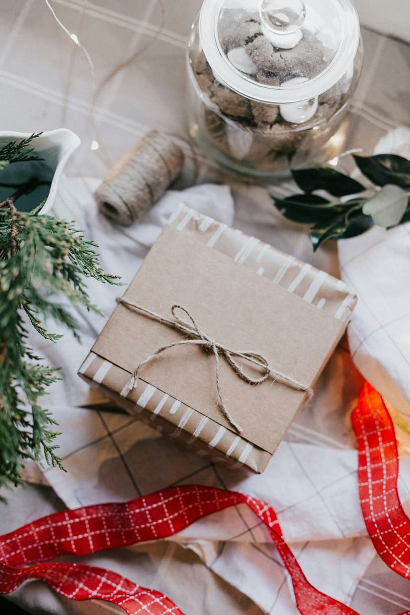 Brown paper gift with twine sits near a cookie jar and red patterned ribbon on a festive holiday tabletop.