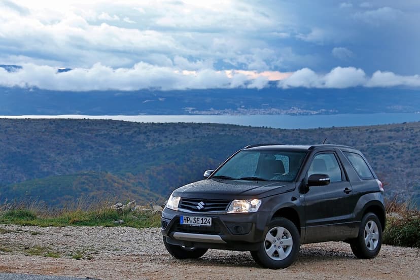 Dark Suzuki Grand Vitara SUV parked on a gravel mountain ridge with a stormy sky and distant sea view.