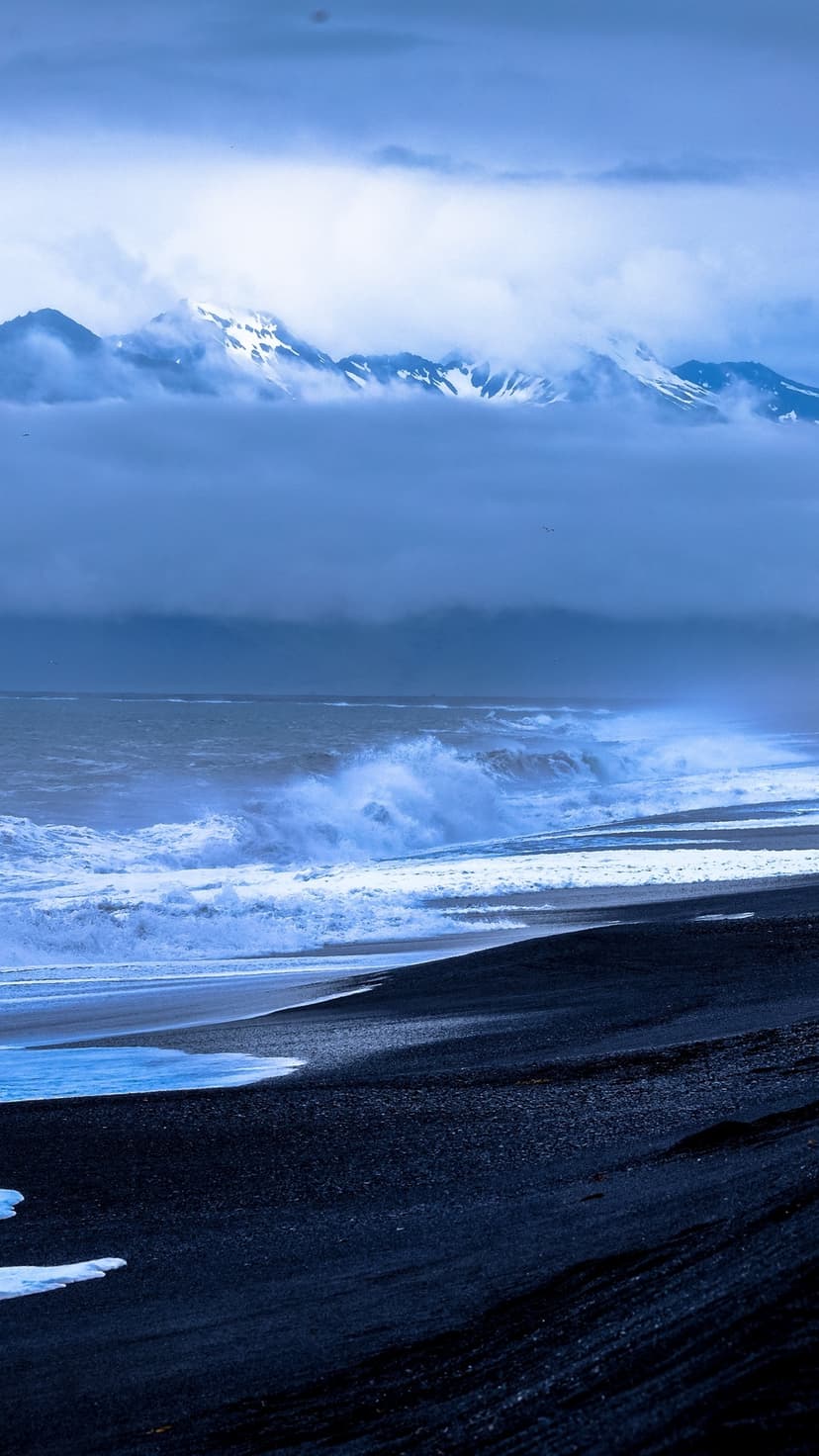 Turquoise ocean waves break on a black volcanic beach with snow-covered mountains under a heavy gray sky.