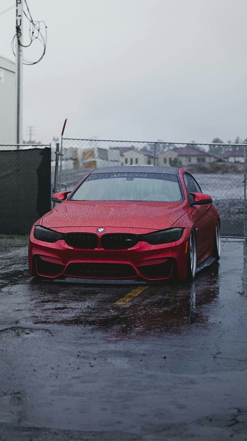 Red BMW M4 coupe parked on wet urban asphalt with rain droplets on its glossy finish and reflections in puddles.