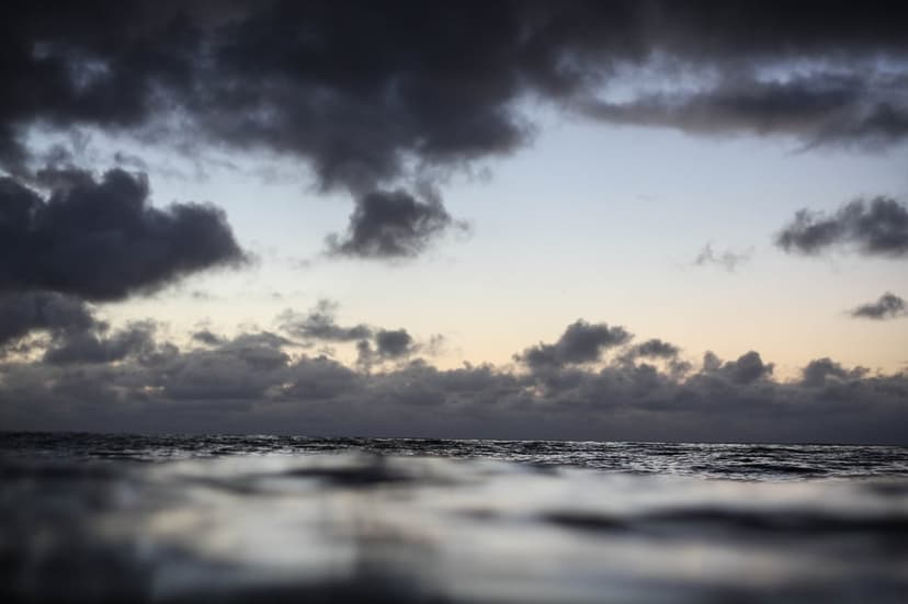 Churning ocean waves under heavy storm clouds at dusk with a dim light glow along the distant horizon line.