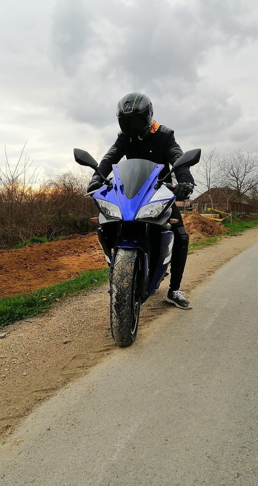 Motorcyclist in a black jacket and helmet sits on a blue and black sportbike on a paved road under clouds.