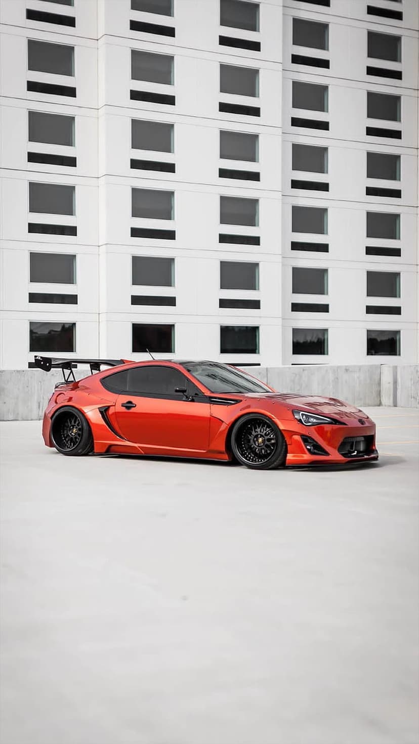 Orange sports car with a large rear spoiler and black wheels parked in front of a white gridded building.