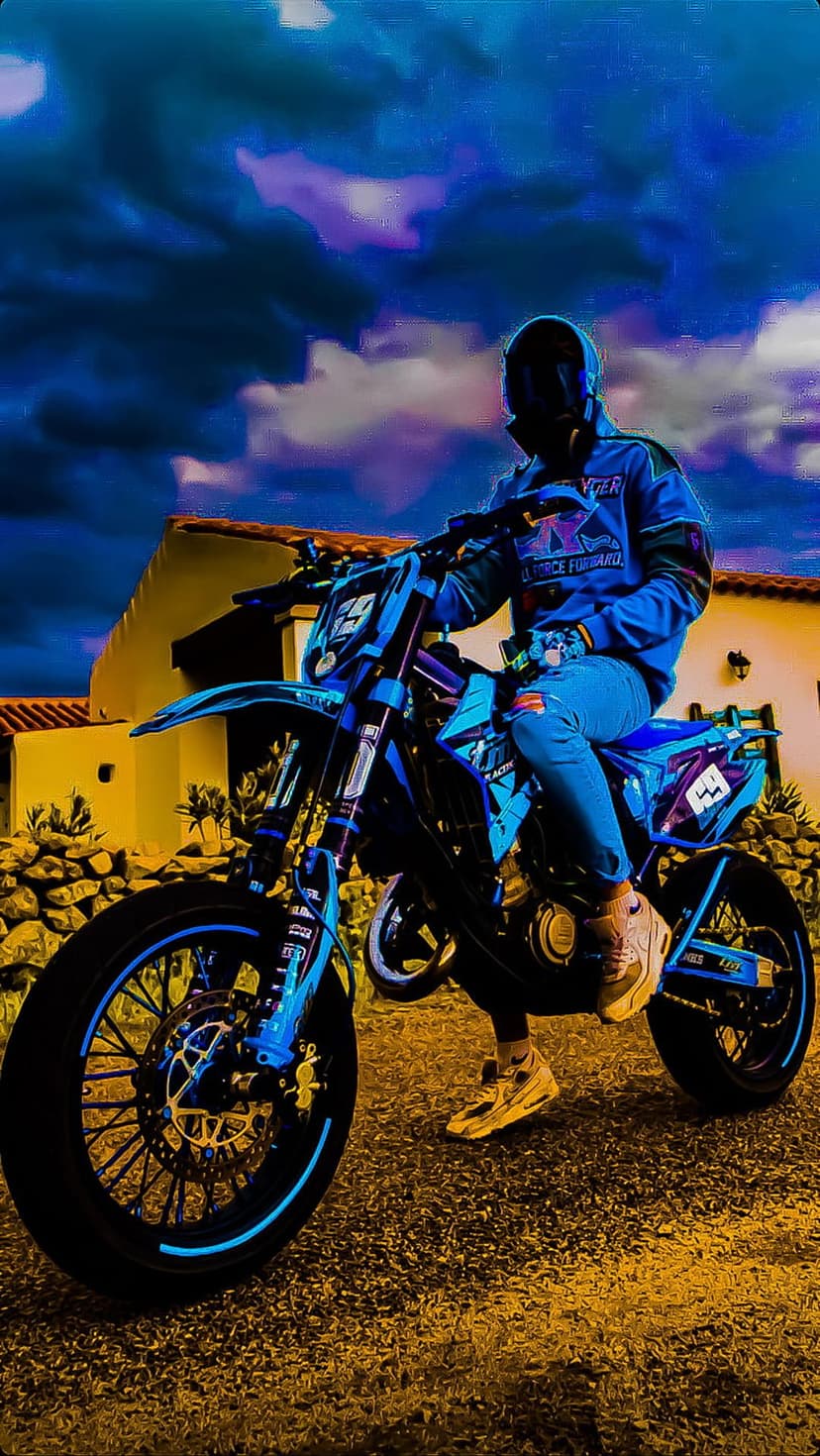 Motocross rider in blue gear sits on a bike under a swirling purple sky beside a rustic yellow building.