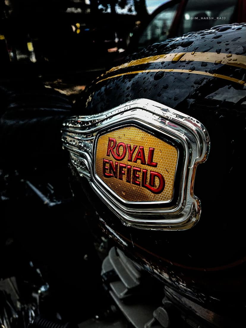 Royal Enfield chrome emblem on a black motorcycle tank covered in raindrops and featuring gold pinstripes.