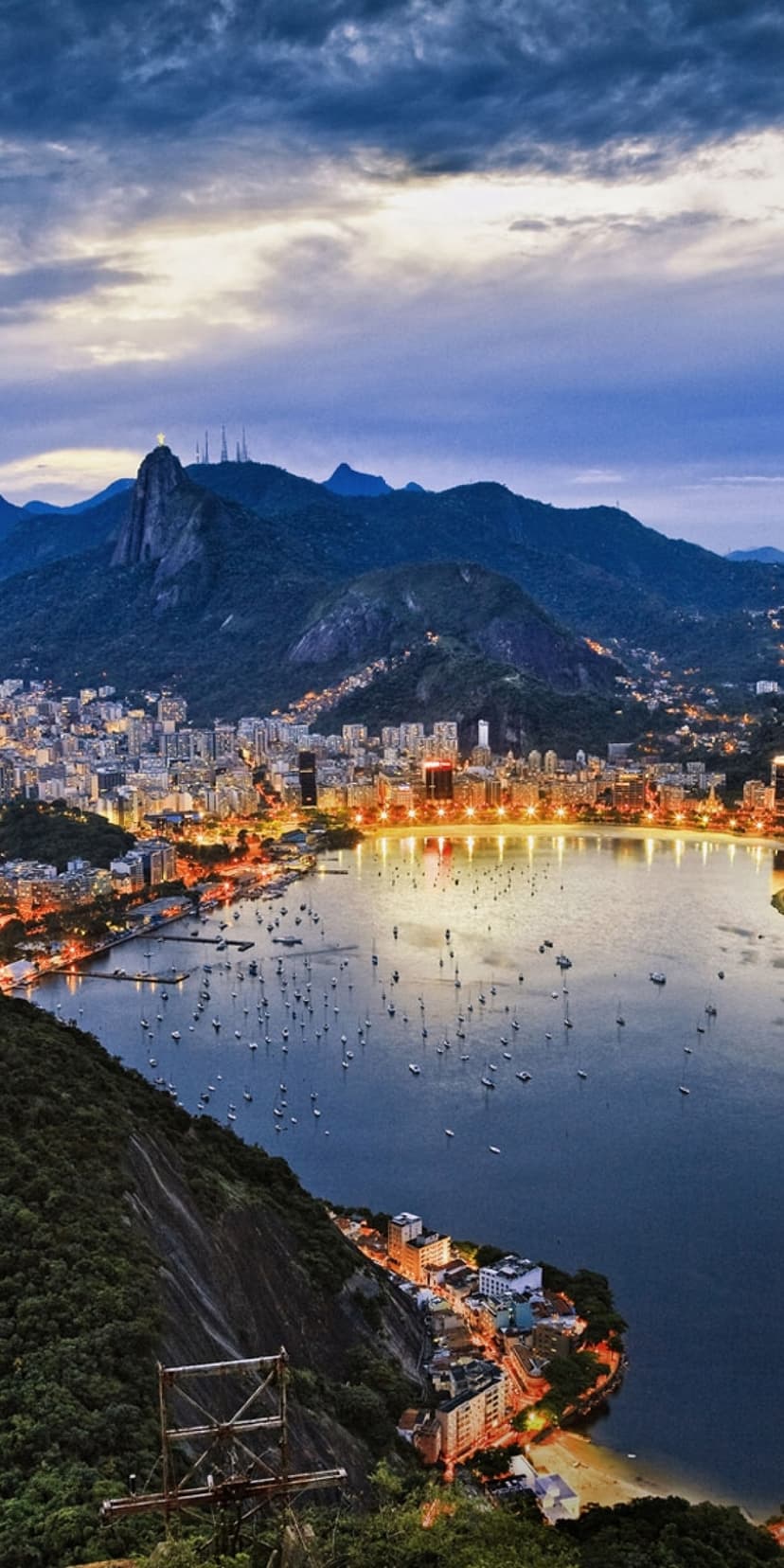 Rio de Janeiro city lights encircling Guanabara Bay at dusk with dark mountains and boats on the water.