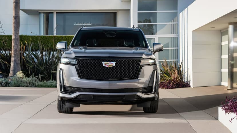 Silver Cadillac Escalade with a black mesh grille parked in front of a white modern architectural home.