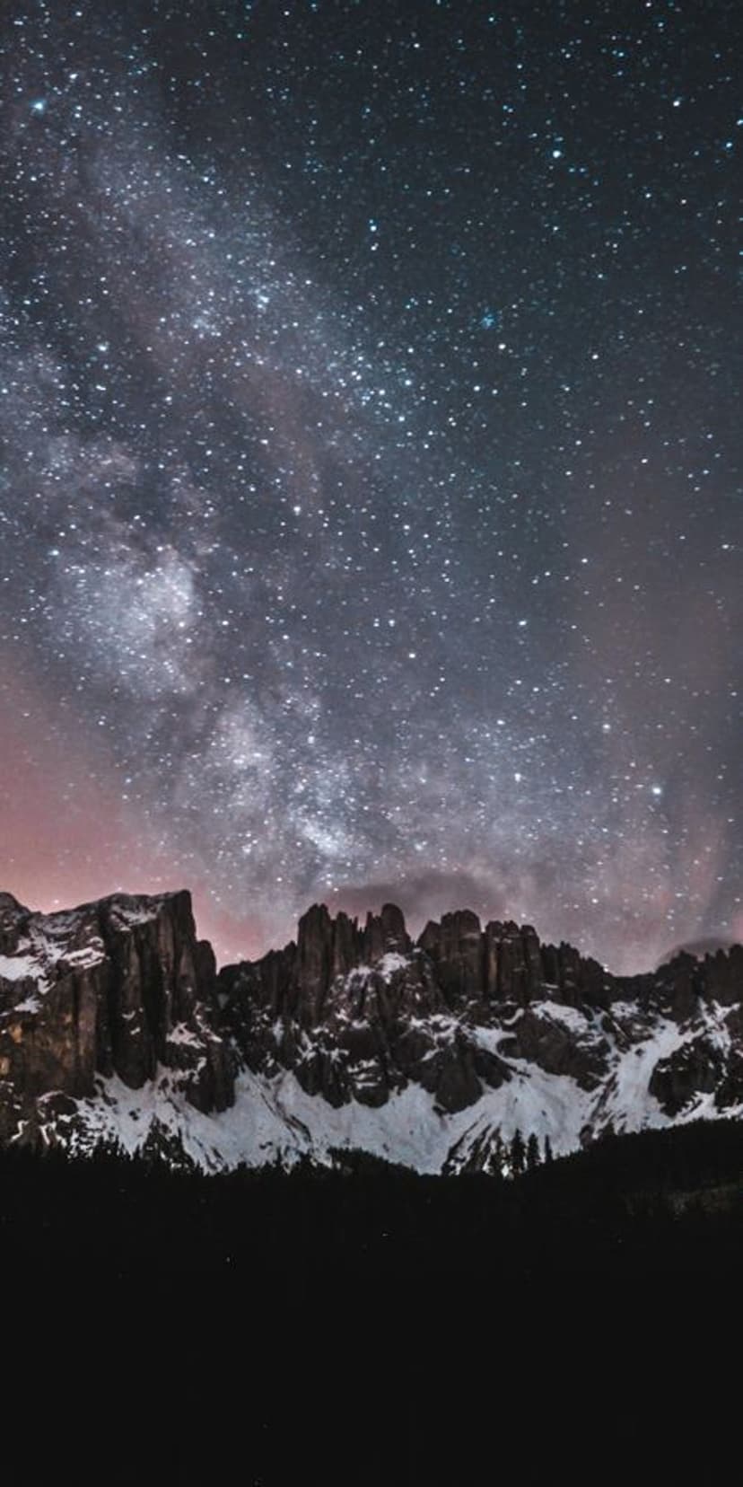 Jagged snow-covered mountain peaks stand beneath a glowing purple and blue Milky Way galaxy in a dark night sky.