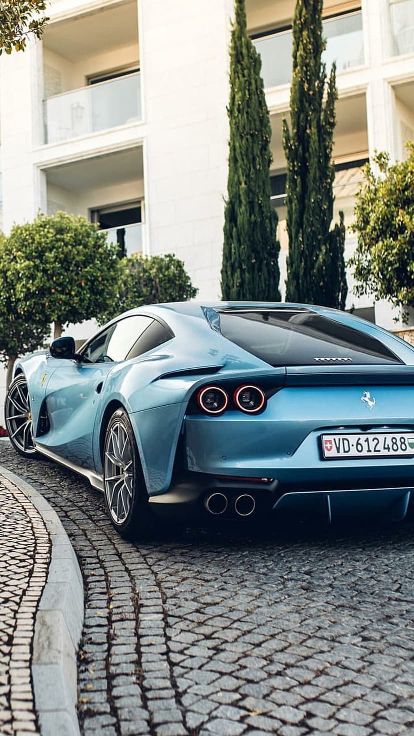Blue Ferrari supercar parked on a cobblestone street with modern white architecture and tall cypress trees.