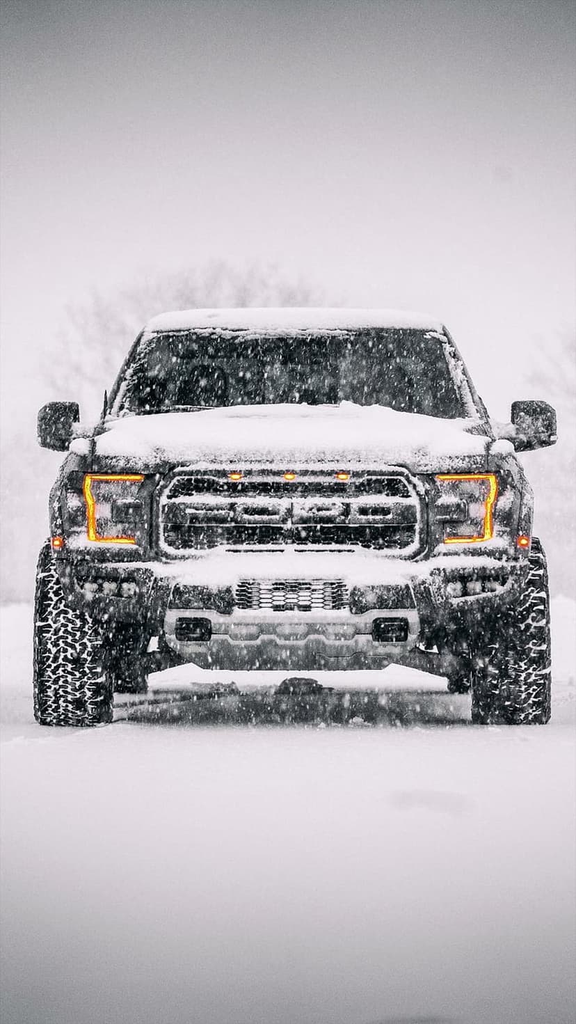 Black Ford Raptor truck driving through a heavy blizzard with glowing headlights and snow-covered off-road tires.