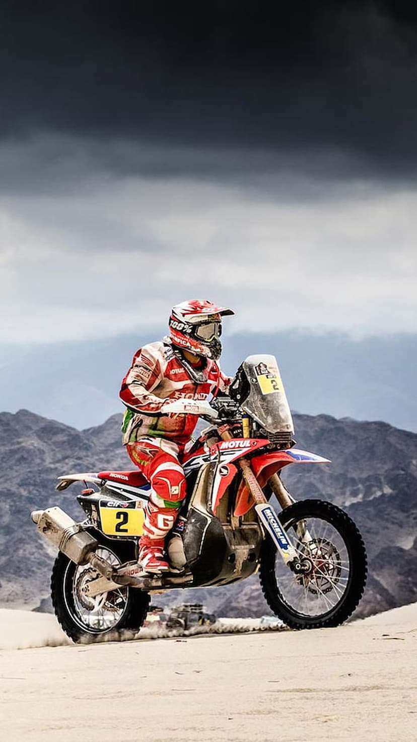 Motorcyclist on a red and white bike number 2 races across sandy dunes against a backdrop of dark mountains.