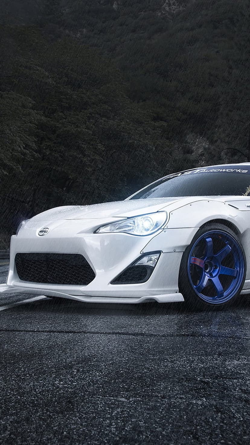 White sports car with widebody fenders and blue wheels parked on a wet asphalt road against a dark hillside.