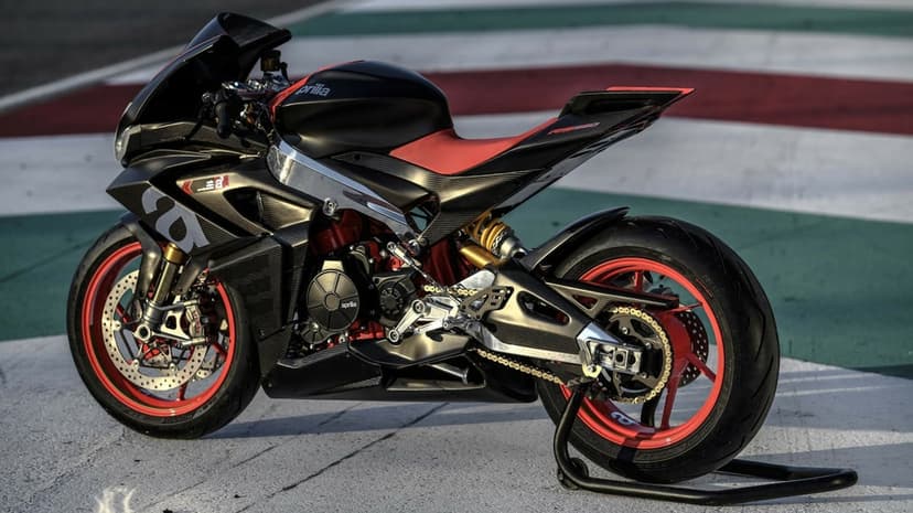 Side view of a carbon fiber Aprilia sportbike with red wheels and seat on a racetrack with Italian flag colors.