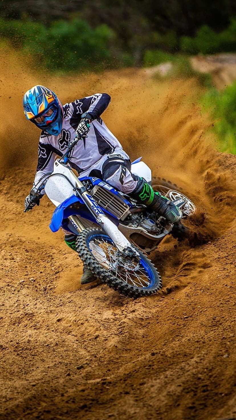 Motocross rider in protective gear leans a blue motorcycle into a sharp turn kicking up orange dust and sand.