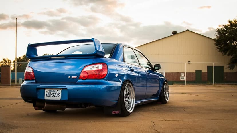 Blue Subaru WRX STI with wide wheels and a large wing parked at sunset near an industrial warehouse building.