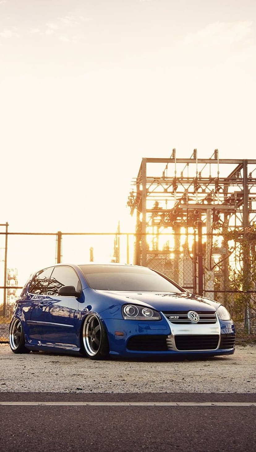 Blue VW Golf R32 with a lowered stance parked on gravel near an industrial substation during golden hour.